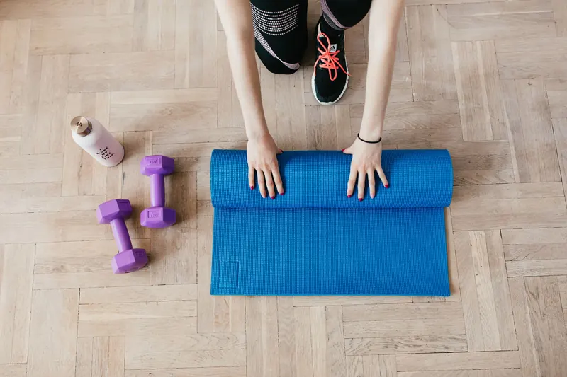 Top view of anonymous female athlete in black leggings and sneakers unfolding blue mat for exercising on floor near dumbbells and water bottle in modern fitness