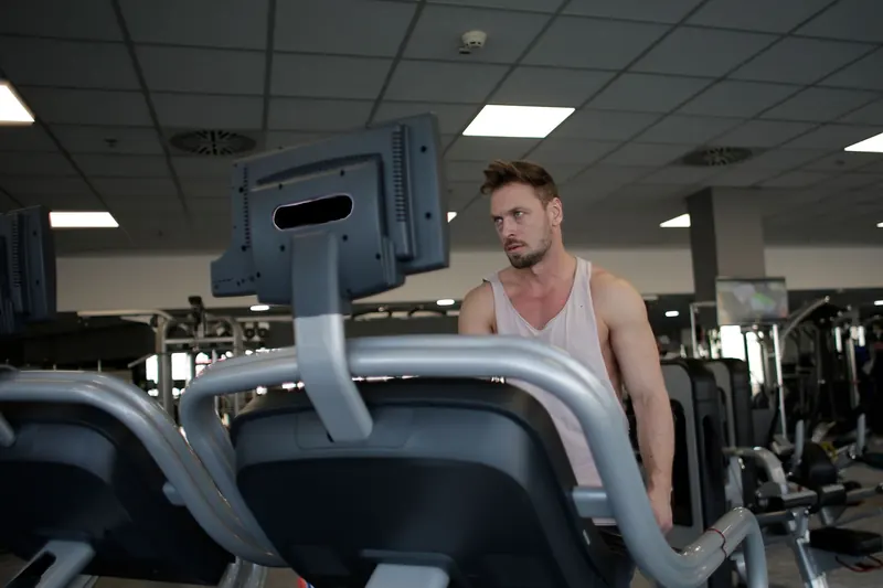Confident muscular adult male athlete in sportswear standing near running machine in modern spacious fitness center before training while looking away pensively