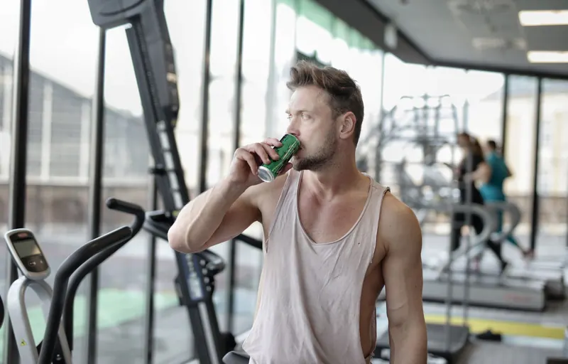 Strong muscular young male athlete in casual sportswear drinking fresh cold energy drink while resting after intense workout and looking away through window in 